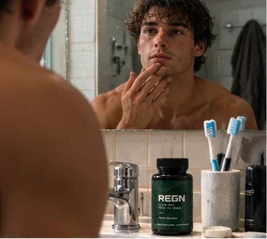 Man checking his skin in the bathroom mirror, REGN bottle by the sink